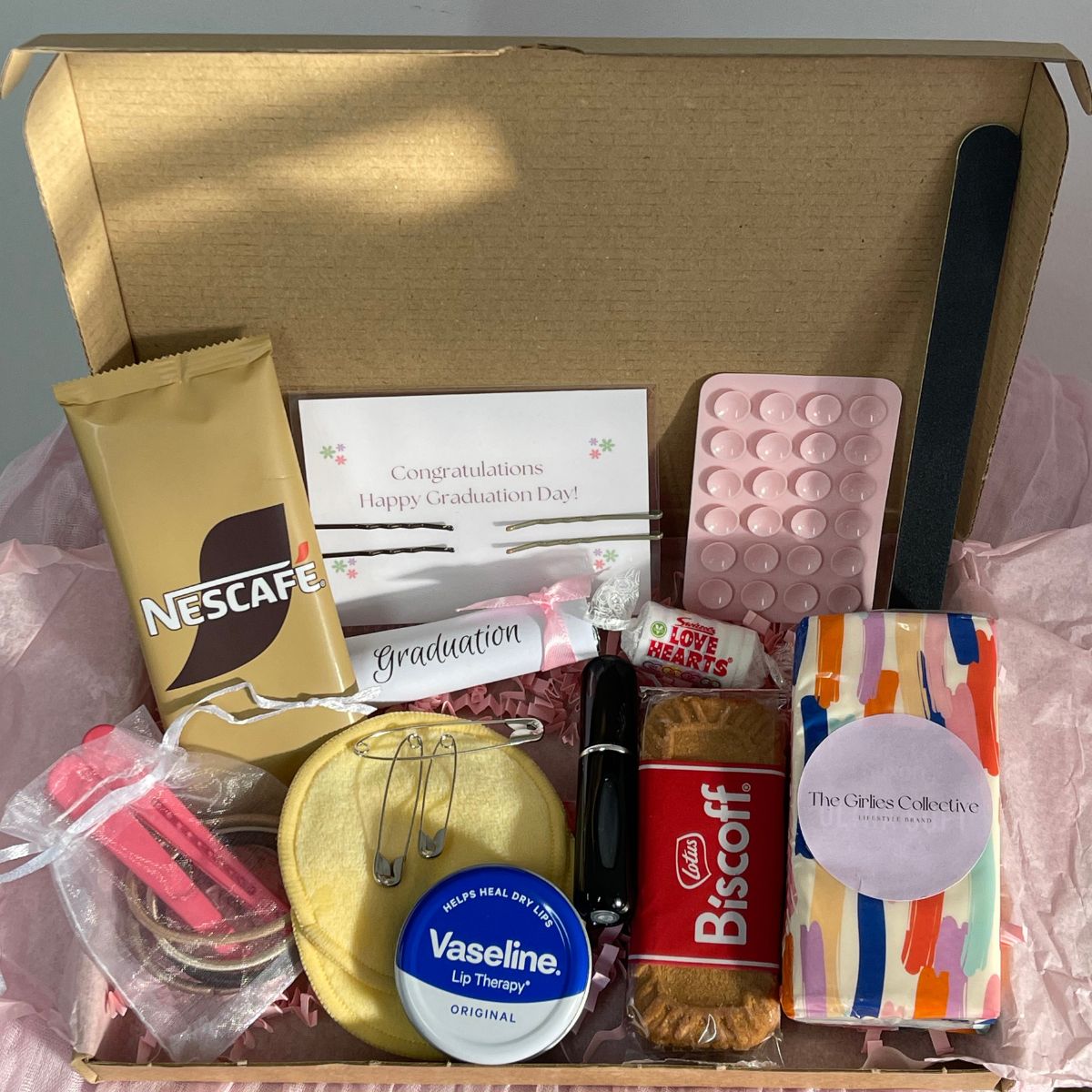Image of 'It's Graduation Day' Self-Care Gift Box which contains: 
Tissues
Vaseline
Perfume Atomiser (perfume not included)
Hairbands/Hair Grips/Hair Slides/Safety Pins
Nail File
Cloth Cleansing Pads
Sticky Phone Mount
Coffee Sachet
Biscoff Biscuit
Love Hearts Sweets
Graduation Polo Mints
PLUS a FREE The Girlies Collective Bookmark!